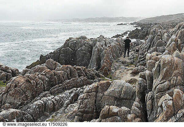Ein Mann geht auf den zerklüfteten Felsen und der felsigen Küste des Atlantiks am Strand von De Kelders spazieren  während sich die Wellen am Ufer brechen.