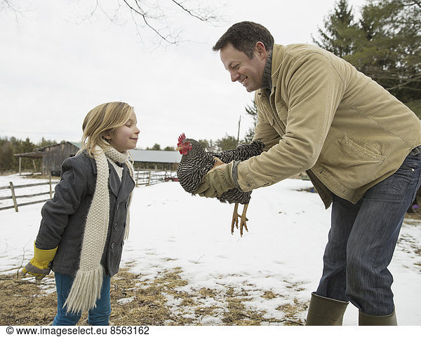 Ein Mann hält ein Huhn in den Händen. Ein Mädchen lehnt sich nach vorne. Ein Bauernhof im Schnee.