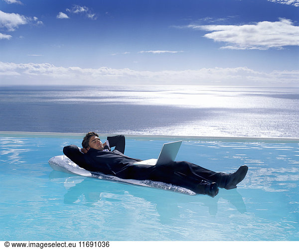 Ein Mann im Anzug auf einer Luftmatratze im Schwimmbad  ein Laptop auf seinem Schoß.