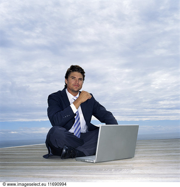 Ein Mann in Anzug und Krawatte sitzt auf einem Deck am Meer und arbeitet an einem Laptop.
