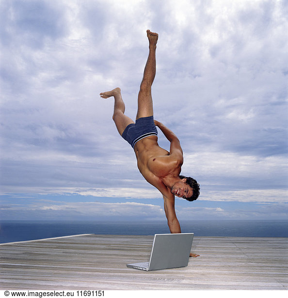 Ein Mann in Badehose macht auf einem Deck am Meer einen einarmigen Handstand  ein Laptop an seiner Seite.