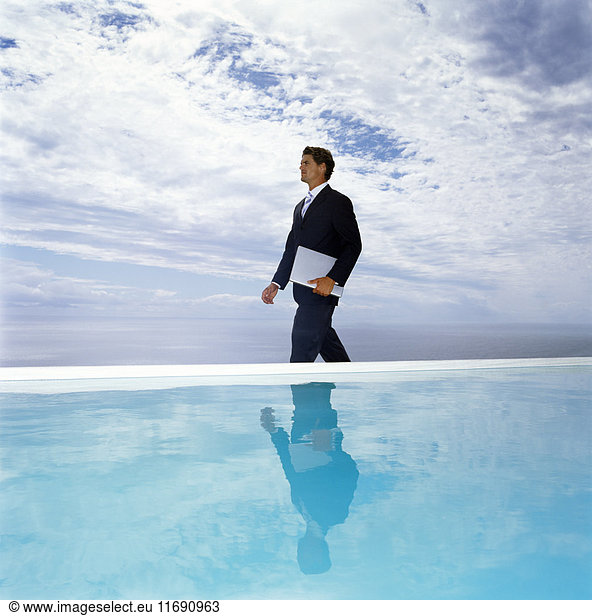 Ein Mann in einem Anzug  der mit einem Laptop an einem Schwimmbad entlang geht.