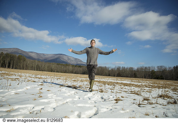 Ein Mann in einem Zopfmusterpullover und Miststiefeln steht mit ausgestreckten Armen in einer verschneiten ländlichen Landschaft.