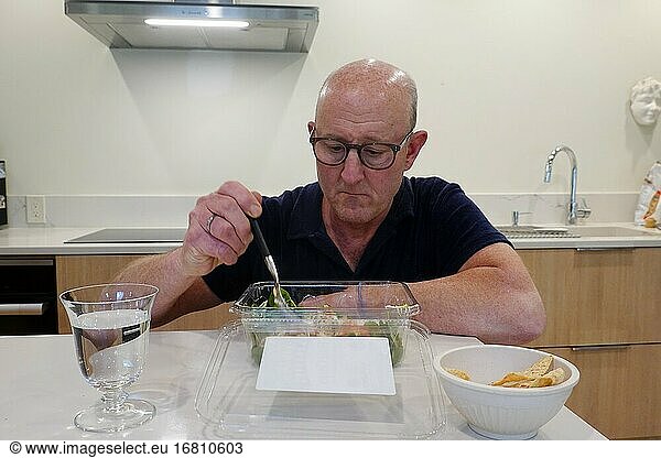 Ein Mann isst allein in einer Küche einen Salat zum Mitnehmen.