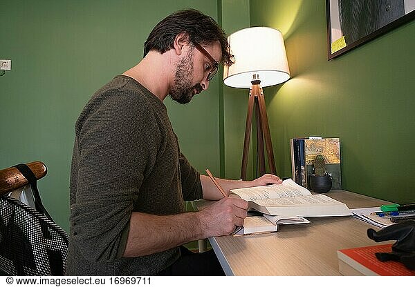 Ein Mann liest in seinem Arbeitszimmer ein Buch und macht sich Notizen  während sein Arm in der Quarantänezeit auf dem Schreibtisch liegt.