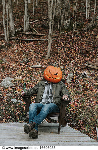 Ein Mann mit einem gruseligen Kürbiskopf sitzt zu Halloween mit einem Bier auf einem Steg.