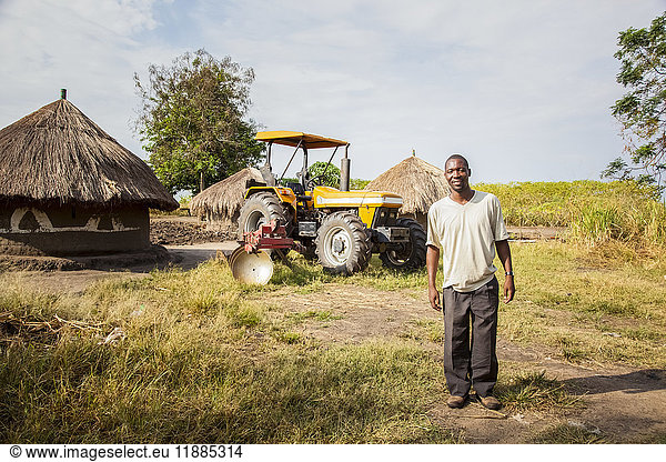 Ein Mann posiert mit einem Traktor und einer Hütte im Hintergrund; Uganda'.