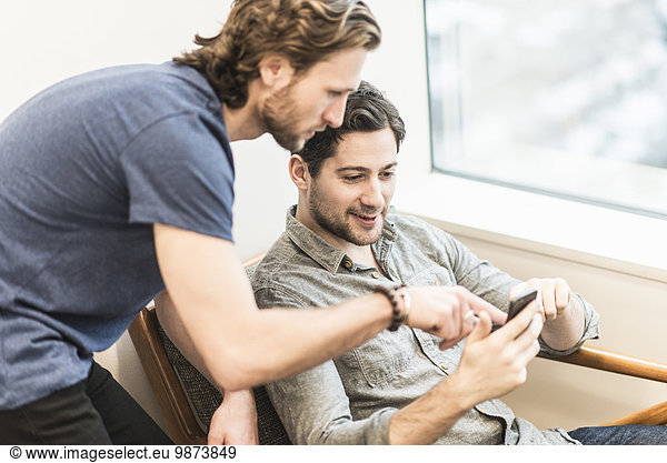 Ein Mann sitzt und schaut auf sein Smartphone und ein Kollege schaut ihm über die Schulter.