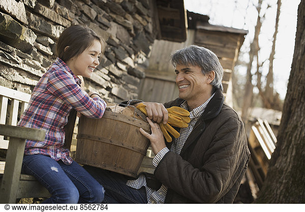 Ein Mann und ein Kind auf einem Biohof. Ein junges Mädchen sitzt auf einem Zaun  mit einem Holzeimer. Ein Mann mit Arbeitshandschuhen.