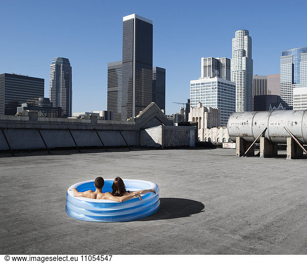 Ein Paar  ein Mann und eine Frau  sitzen in einem kleinen aufblasbaren Wasserbecken auf dem Dach einer Stadt und kühlen sich ab.