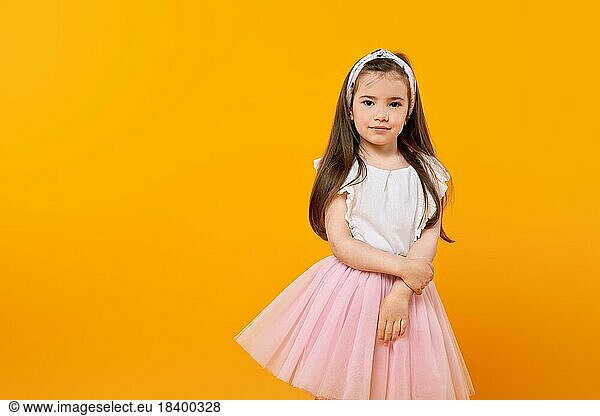 Ein süßes kleines Mädchen steht in einem gelb getönten Studio und schaut unschuldig in die Kamera. Ihr langes Haar und ihr modisch geschmücktes Kleid spiegeln ihre Kindheit wider  während sie diesen Moment der Schönheit festhält