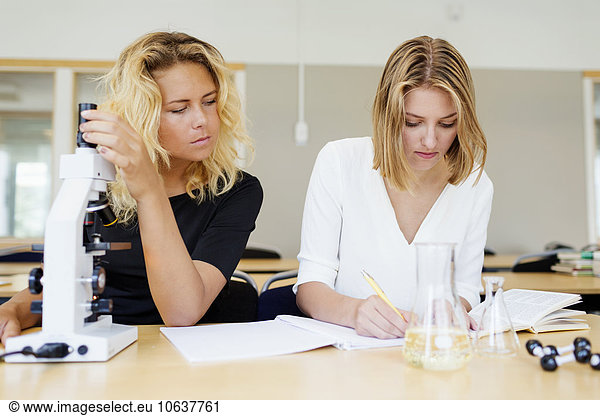 Ein Student der Naturwissenschaften benutzt ein Mikroskop  während ein Mitschüler im Labor in ein Buch schreibt