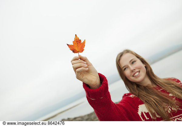 Ein Tagesausflug am Ashokan-See. Ein junges Mädchen in einem roten Winter-Strickpullover  das ein Ahornblatt hochhält.