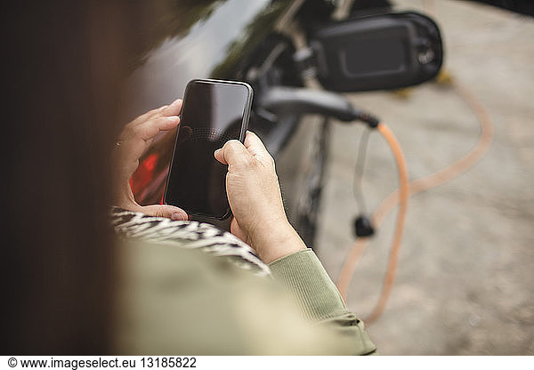 Ein Teil der Frau benutzt ein Smartphone  während sie im Stehen ein Elektroauto auflädt