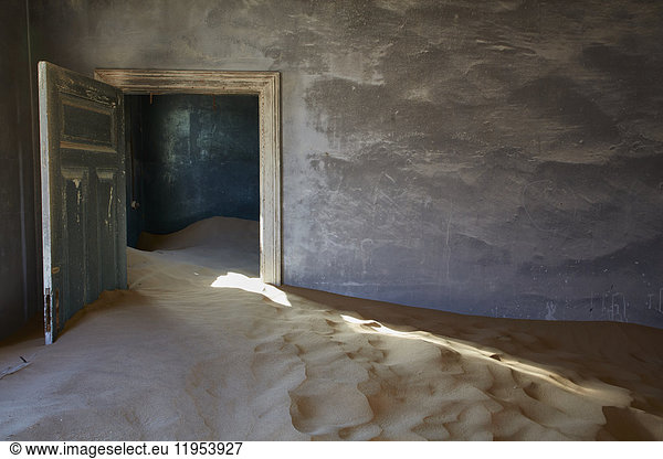 Ein verlassenes Gebäude  eine offene Tür und Sandverwehungen  die vom Rest des Gebäudes in den Raum eindringen. Eine Geisterstadt.