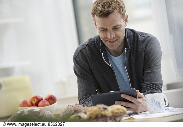 Eine Büro- oder Wohnungseinrichtung in New York City. Ein Mann in einem Sweatshirt-Oberteil mit einem digitalen Block. Er lehnt sich an eine Frühstückstheke.