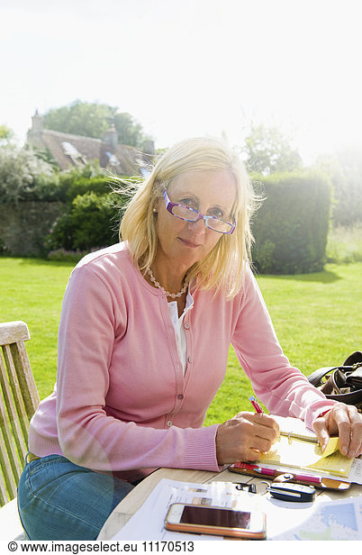 Eine blonde Frau  die in einem Garten an einem Tisch sitzt  mit einem Smartphone und einem Notebook  die im Freien arbeitet.