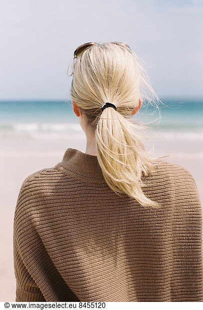 Eine blonde Frau mit blonden Haaren  die von der Küste aufs Meer hinaussieht.
