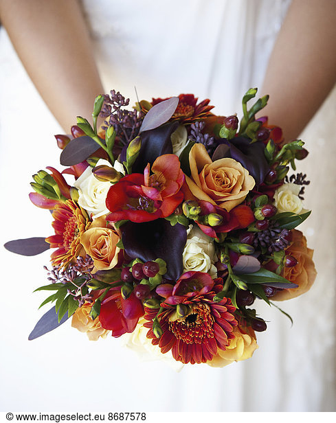 Eine Braut hält einen Brautstrauß aus bunten roten und orangefarbenen Blumen mit violett getönten Blättern.