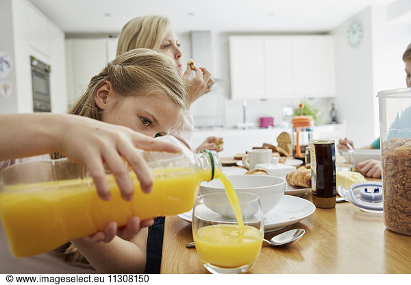 Eine Familie sitzt beim Frühstück  ein Mädchen schenkt ein Glas Orangensaft ein.