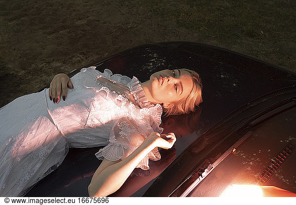 eine Frau in einem Kleid  die abends auf der Motorhaube eines Autos liegt