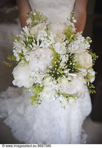 Eine Frau in einem weißen Kleid  eine Braut mit einem Brautstrauß aus weißen Blumen  großen weißen Rosen und Pfingstrosen  mit zarten gelben Blüten und grünen Blättern.
