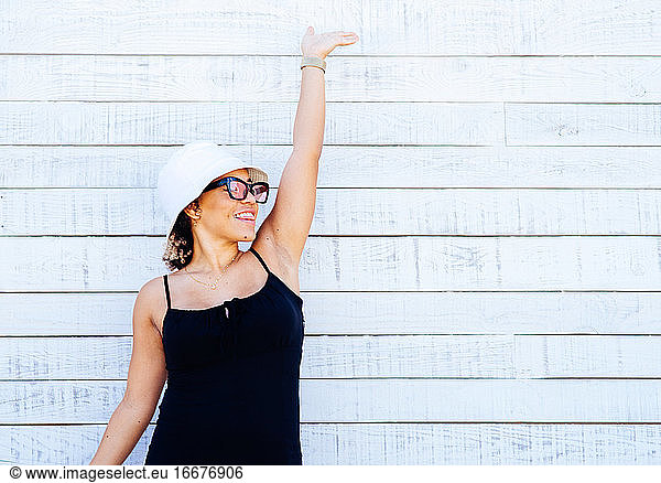 Eine Frau mit weißem Hut und Sonnenbrille steht an einer weißen Wand.