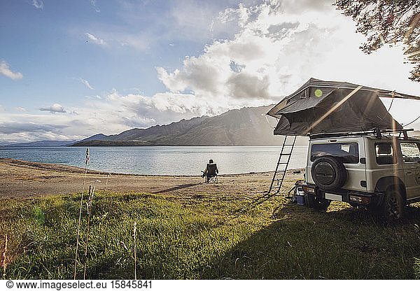 Eine Frau sitzt in Neuseeland an einem See neben einem Geländewagen mit einem Zelt