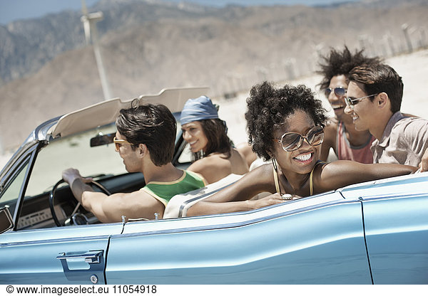 Eine Gruppe von Freunden in einem hellblauen Cabriolet auf offener Straße  die über eine trockene  flache  von Bergen umgebene Ebene fahren.