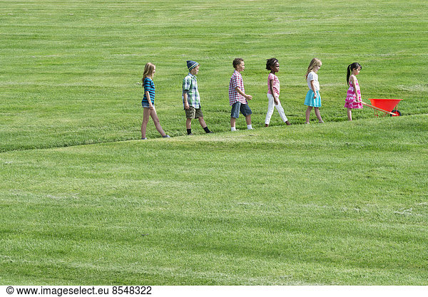 Eine Gruppe von Kindern  die einen schrägen Weg in Höhenrichtung hinaufgehen und dem kleinsten vorne mit einer roten Schubkarre folgen.