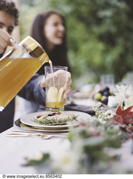 Eine Gruppe von Menschen um einen Tisch in einem Garten. Ein Festmahl mit gedecktem Tisch und Blattdekoration. Eine Person  die Getränke in Gläser gießt.