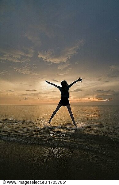 Eine junge Frau macht einen Sternsprung und genießt ihre Freiheit am Strand bei Sonnenuntergang.