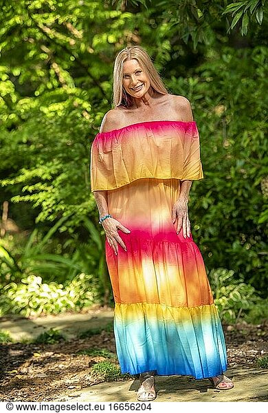Eine lächelnde 51-jährige blonde Frau in einem farbenfrohen Kleid schaut in die Kamera  draußen in einer Gartenanlage.