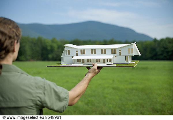Eine ländliche Szene und eine Bergkette sowie eine Person  die ein maßstabsgetreues Modell eines neuen Gebäudes hält.