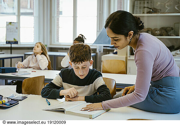 Eine Lehrerin hilft einem Schüler beim Lesen in einem Buch  während er auf dem Schreibtisch im Klassenzimmer sitzt