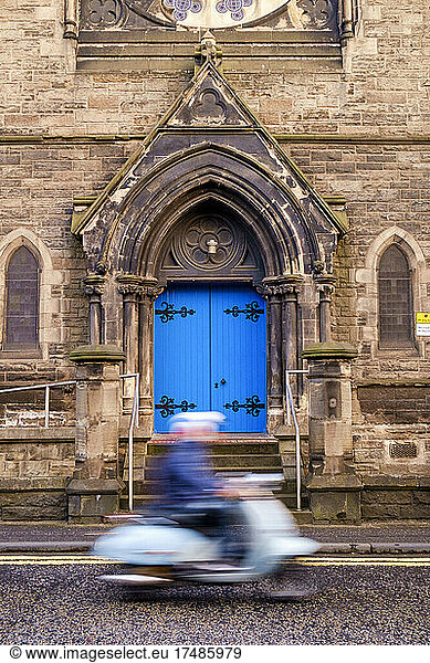 Eine Person auf einem Motorroller fährt an einem gotischen Bogen mit hellblauer Tür vorbei.