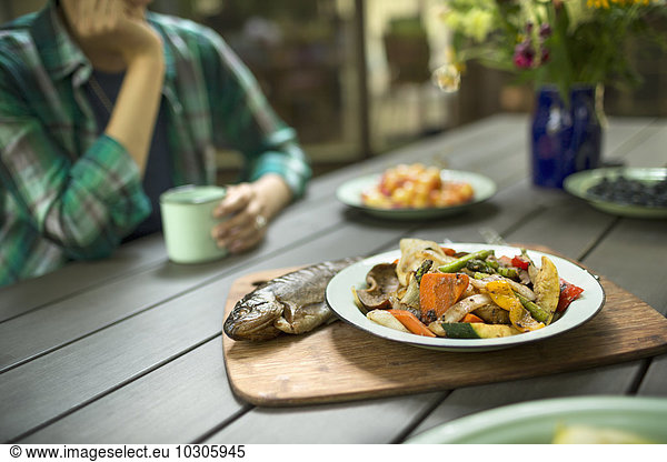 Eine Person  die an einem Tisch im Freien sitzt. Ein gekochter frischer Fisch und ein Gericht mit Gemüse.