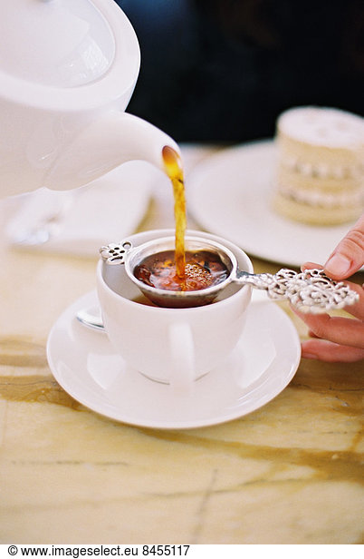 Eine Person  die mit einem Sieb eine Tasse Tee einschenkt. Weißes Porzellan. Eleganter Nachmittagstee.