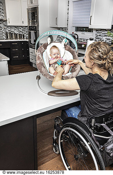 Eine querschnittsgelähmte Mutter im Rollstuhl  die mit ihrem Baby im Kindersitz spielt  während es in ihrer Küche sitzt; Edmonton  Alberta  Kanada