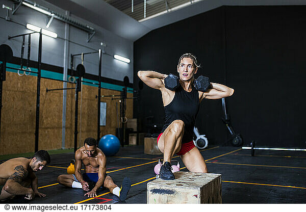 Eine Sportlerin hebt Hanteln  während sie auf einer Box trainiert  und ein Mann ruht sich im Fitnessstudio aus.