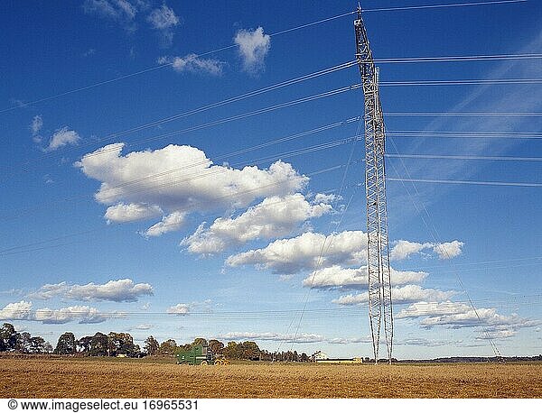 Eine Stromleitung quer über das Haferfeld mit einem Mähdrescher und weißen Wolken am blauen Himmel. Hinterland. Schweden.