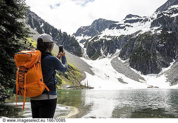 Eine Wanderin mit Rucksack und Smartphone steht in der Nähe eines Sees und fotografiert die malerische Landschaft der schneebedeckten felsigen Berge während einer Sommerreise in British Columbia