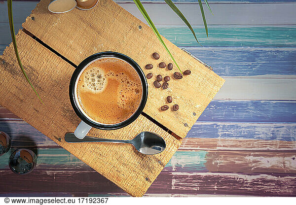 Eine weiße Kaffeetasse mit Sahnekaffee steht auf einer farbigen Holzfläche. Kaffeebohnen stehen daneben