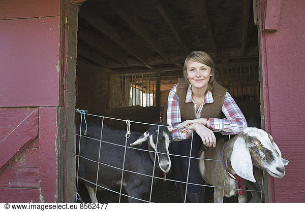 Eine Ziegenfarm. Ein junges Mädchen lehnt an der Absperrung des Ziegenstalls  aus dem zwei Tiere herausschauen.