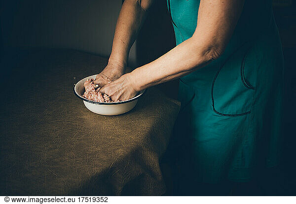 Elderly woman kneads minced meat in bowl on kitchen table