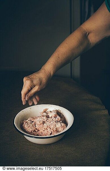 Elderly woman salts minced meat in bowl on kitchen table