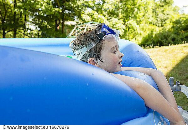 Elementary Age Boy Wearing Goggles on Forehead Relaxes on Side of Pool