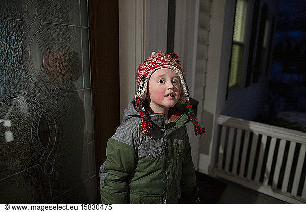 Elementary age boy wears cold weather gear standing at night on porch