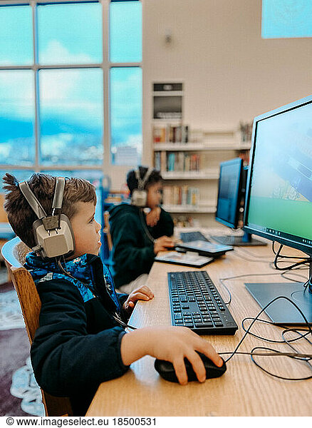 Elementary brothers playing on computers at library with headphones