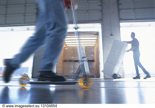 Employee on a push along scooter moving through a large warehouse  and a man stacking large boxes for dispatch in a distribution warehouse loading dock area.